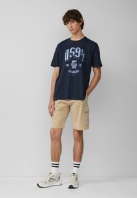 Marineblauwe katoenen T-shirt met een grafische print in lichterblauw, gecombineerd met khaki cargoshorts en witte sneakers met grijze accenten en zwart gestreepte sokken.
