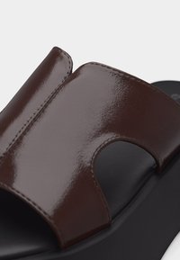 Λουρί σανδαλιού από σκούρο καφέ γυαλιστερό δέρμα με σχέδιο κοψίματος πάνω σε μαύρη μαλακή σόλα.