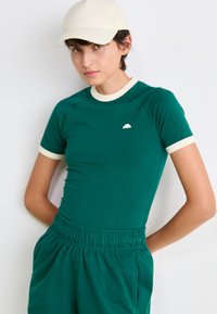 Grøn fitted t-shirt med cremefarvet kant omkring halsen og ærmerne, med et lille hvidt logo. Sammen med matchende grønne shorts med elastik i taljen.