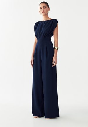 Donna con capelli raccolti indossa una tuta senza maniche blu navy a gamba larga e un bracciale rigido dorato, in piedi davanti a uno sfondo bianco.