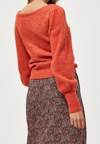 Oranger Strickpullover mit weitem Ausschnitt und langen, leicht gepufften Ärmeln, kombiniert mit einem floral gemusterten Rock in Rot- und Weißtönen.