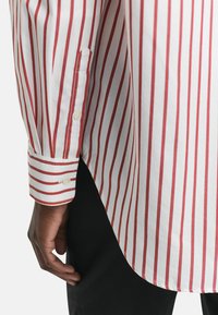 White button-up shirt with narrow red stripes. Features classic collar, single-button cuffs, and a high-low hem design. Smooth cotton texture.