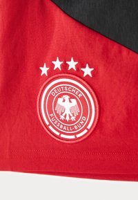 Κόκκινη αθλητική φανέλα με κεντητό διακριτικό σε κυκλικό σχήμα με σχέδιο αετού, πέντε λευκά αστέρια από πάνω και κείμενο "DEUTSCHER FUSSBALL-BUND".