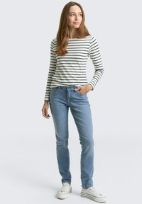 Kobieta stoi z jedną ręką w kieszeni jasnoniebieskich dżinsów, ubrana w białą koszulę w czarne paski z długim rękawem oraz białe trampki.