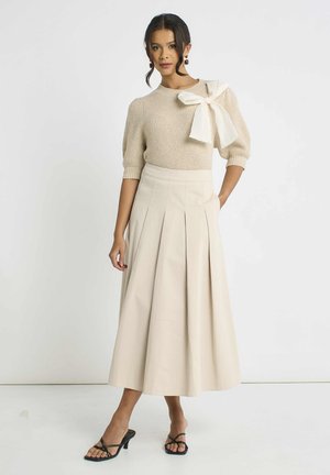 Woman wearing beige knit sweater with large white bow on shoulder, cream pleated midi skirt, and black heeled sandals, standing against white background.