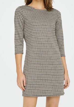 Frau trägt ein eng anliegendes, beige kariertes Kleid mit dreiviertel Ärmeln und einem runden Ausschnitt, stehend vor einem schlichten Hintergrund.