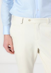 Cremefarbene maßgeschneiderte Hosen mit flachem Frontdesign, seitlichen Taschen und einem einzelnen Knopfverschluss. Das dazu passende Hemd ist hellblau und hat eine glatte Textur.