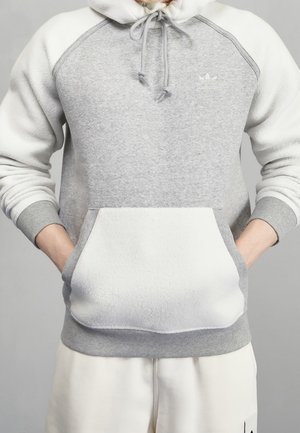 Henkilö pukeutuneena harmaavalkoiseen Adidas-huppariin, kädet etutaskussa ja valkoiset housut yksiväristä harmaata taustaa vasten.