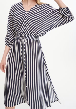 Donna che indossa un vestito a righe blu navy e bianche con maniche a pipistrello e cintura in vita, in piedi davanti a uno sfondo bianco neutro.