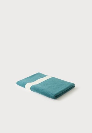 Serviette bleu sarcelle pliée avec une large bande blanche au centre sur un fond clair uni.