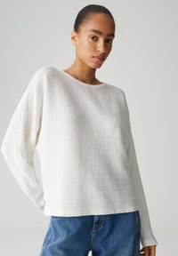 Maglione bianco a maniche lunghe con trama, scollo ampio e spalle scese, abbinato a jeans di denim blu. Tessuto liscio, vestibilità ampia.