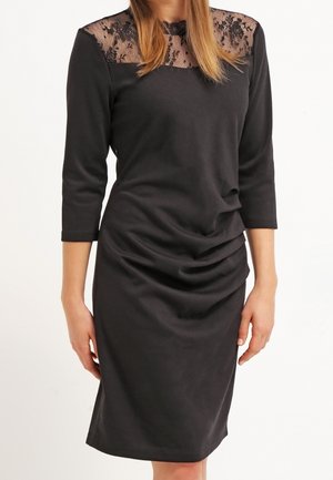 Femme portant une robe noire longueur genou avec des manches trois-quarts et un empiècement en dentelle transparente à motifs floraux.