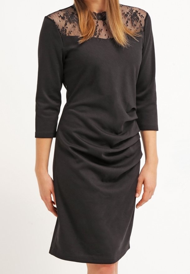 Femme portant une robe noire longueur genou avec des manches trois-quarts et un empiècement en dentelle transparente à motifs floraux.