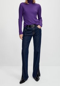 Paarse geribbelde coltrui en donkerblauwe wijde jeans met spleten aan de zijkant. Zwarte riem met zilverkleurige gesp. Gladde texturen en een aansluitend ontwerp.