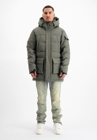 Man die staat in een olijfgroene gewatteerde jas met grote zakken, lichte vervaagde jeans en witte sneakers tegen een witte achtergrond.