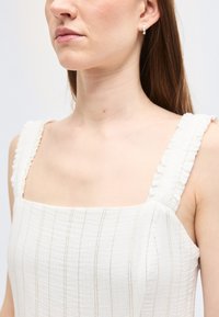 Γυναίκα που φοράει λευκό αμάνικο φόρεμα με κάθετες ρίγες και βολάν στο τιράντες, εμφανίζεται από τους ώμους έως το πηγούνι, κοιτάζοντας προς τα αριστερά.