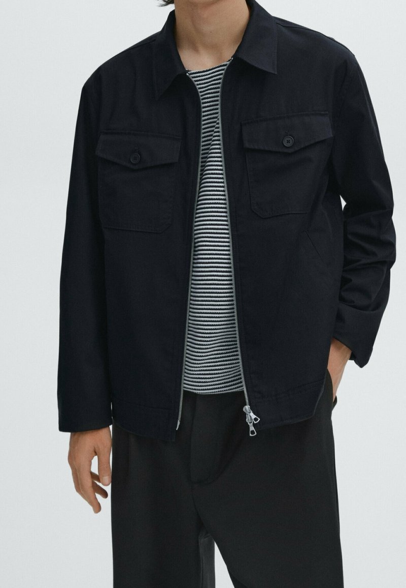 Homme portant une veste noire zippée par-dessus une chemise rayée noir et blanc avec un pantalon noir, une main dans la poche et une autre le long du corps.