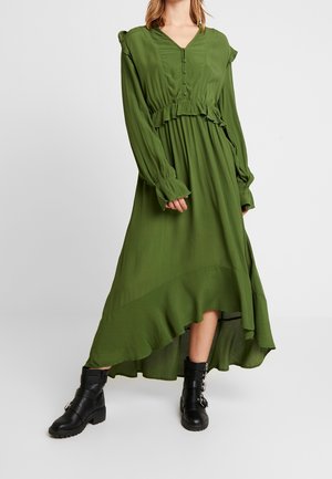 Grünes, langärmliges Kleid mit V-Ausschnitt, gerafftem Taillendetail und asymmetrischem Saum; kombiniert mit schwarzen Stiefeletten. Weiche Stofftextur.