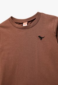 Brązowa bawełniana koszulka z okrągłym dekoltem i krótkimi rękawami. Posiada czarny haftowany wzór dinozaura na lewej stronie klatki piersiowej.