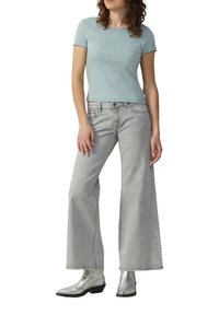 Frau in einem hellblauen Kurzarmoberteil, weit geschnittenen grauen Jeans und metallisch silbernen Stiefeletten, die vor einem weißen Hintergrund steht.