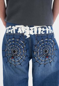 Dżinsy z wyszukanym haftem w kształcie pajęczyny na tylnej kieszeni, z czarnym konturem, czerwonym akcentem i jasnoniebieskim odcieniem.