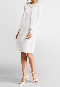 Bílé šaty s krajkovým výstřihem, doplněné bílým svetrem se zapínáním na knoflíky a volánovým límečkem. Outfit je dokončen bílými sandály na podpatku.