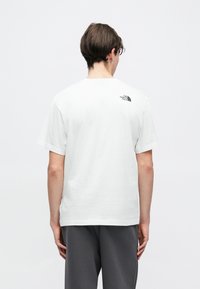 Λευκό βαμβακερό T-shirt με κοντά μανίκια, που διαθέτει μικρό μαύρο λογότυπο στο άνω μέρος της πλάτης. Λείο υφή, σχεδιασμός με κανονικό στρογγυλό λαιμό.