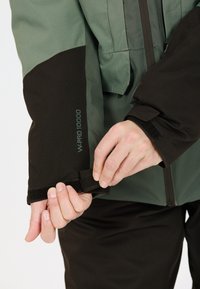Grüne und schwarze wasserdichte Jacke mit verstellbarem Velcro-Aufsatz am Ärmel. Mit einem Reißverschluss vorne und W-PRO 10000-Logo auf dem Ärmel. Strapazierfähige Stofftextur.