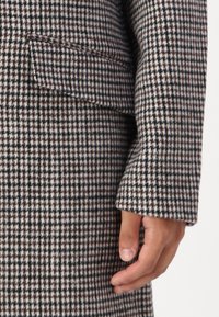 Abrigo de tejido texturizado con patrón de pata de gallo en marrón y beige, que presenta solapa con muesca y bolsillo con solapa. Mano visible descansando sobre el abrigo.