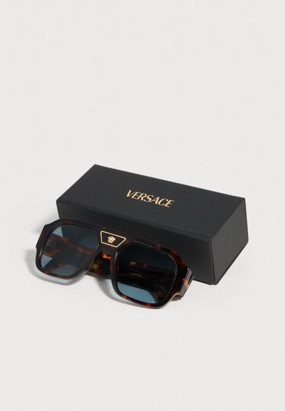 Γυαλιά ηλίου σε σχήμα τετραγώνου με σχέδιο χελώνας και σκούρους φακούς, που ακουμπούν μπροστά από μια μαύρη θήκη με το χρυσό λογότυπο Versace.