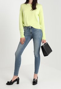 Ljusgrön långärmad topp, blå skinny jeans, svarta högklackade skor och en liten svart handväska med ett metallhandtag.