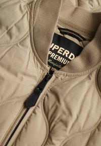 Beige gevoerde jas met een geribde kraag, voorzien van een zwarte ritssluiting en label. Het materiaal heeft een gladde, zachte textuur met een subtiele glans.