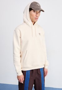 Sweatshirt creme com bolso canguru e capuz com cordão, apresentando um pequeno logotipo castanho da Adidas no peito.
