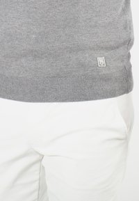 Pull en tricot gris avec ourlet côtelé, doté d'une petite étiquette de logo. Associé à un pantalon blanc pour le contraste. Texture lisse, design décontracté.