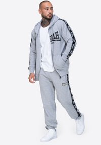 Sudadera gris con cremallera y logo negro, combinada con joggers grises a juego que tienen rayas laterales y zapatillas blancas. Textura de tela suave.