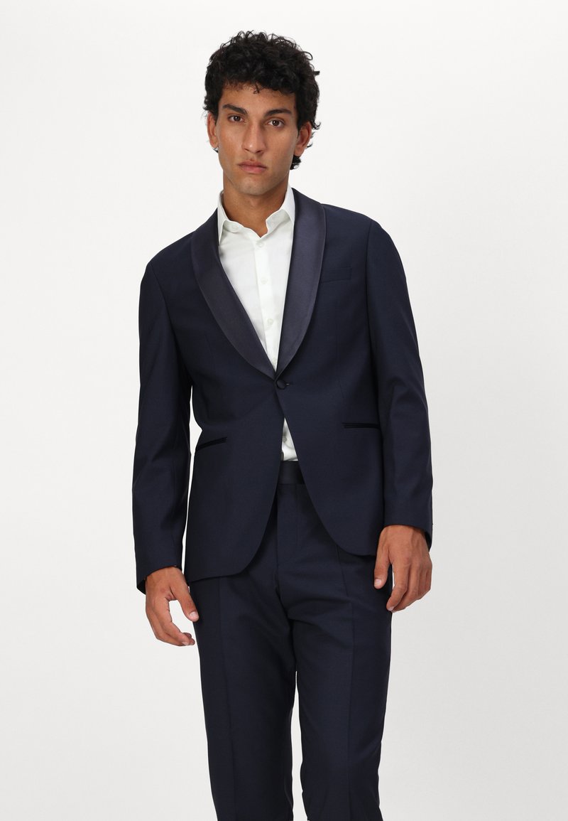 Costume marine avec revers en satin, fermeture à un seul bouton, coupe ajustée et pantalon assorti, portant une chemise blanche à manches longues en dessous.
