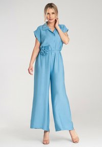 Jasnoniebieski kombinezon z krótkimi rękawami, zapinany na guziki, z materiałowym kwiatem na talii. Miękka tekstura z szerokimi, luźnymi nogawkami.