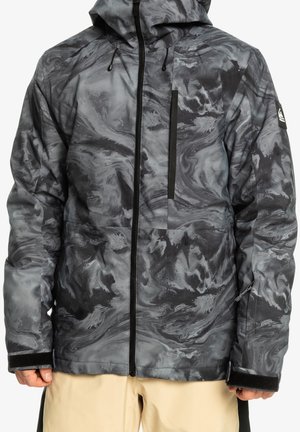 Veste imperméable marbrée gris et noir avec fermeture éclair, capuche, poche poitrine et sangles en velcro aux poignets, portée avec un pantalon beige.
