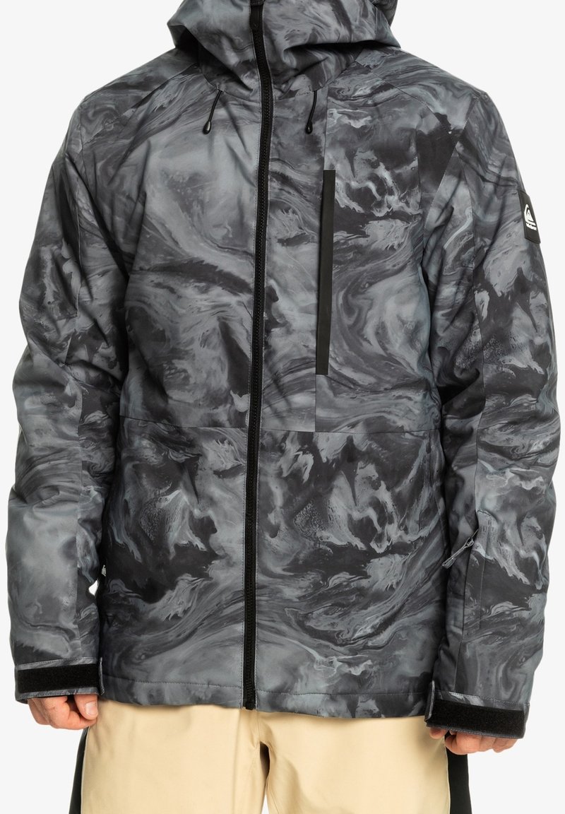 Veste imperméable marbrée gris et noir avec fermeture éclair, capuche, poche poitrine et sangles en velcro aux poignets, portée avec un pantalon beige.