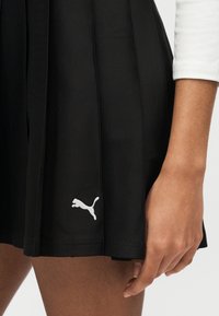 Main d'une personne à côté d'une jupe plissée noire avec un logo Puma blanc près de l'ourlet, portant un haut blanc à manches longues dont la manche s'arrête au-dessus du poignet.