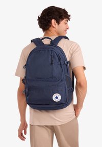 Navyblå ryggsäck med flera fack, med en rund Converse-logotyp och justerbara vadderade remmar för komfort.