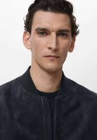 Chaqueta bomber de ante negro con cuello acanalado y cremallera plateada. La tela tiene una textura suave y presenta elementos de diseño minimalistas.