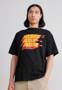 Black cotton t-shirt with oversized fit features yellow and red graphic text design on front. Relaxed sleeves and crew neckline.