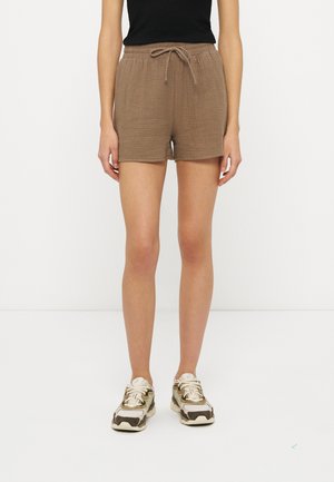 Nő barna, texturált zsinóros rövidnadrágban, fekete felsőben és fehér-beige tornacipőben, fehér háttér előtt állva.