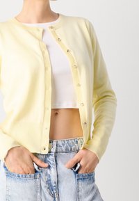 Lichtgele cardigan met een ronde halslijn, knopen aan de voorkant, geribbelde manchetten en zakken, gedragen met een witte crop top en denim shorts.