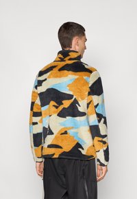 Pullover in pile colorato con un motivo camuffato astratto in arancione, nero, blu e crema. Collo alto e superficie testurizzata.