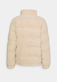 Beige jakke med syntetisk pels og højtstående krave, der har en tekstureret, blød overflade samt vandrette syninger for form og varme.