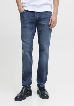 Homme portant un jean bleu foncé, des chaussures habillées noires et une chemise blanche, une main dans la poche, sur un fond uni.