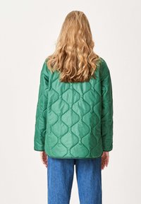 Quiltet grøn jakke med et geometrisk mønster, der har en afslappet pasform, runde kanter og lange ærmer. Båret over blå jeans.