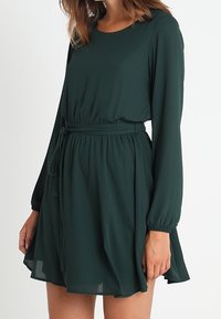 Grünes Chiffonkleid mit langen Ärmeln, tailliertem Bund und fließendem Rock. Mit Gürtel und rundem Ausschnitt. Glatte Textur.